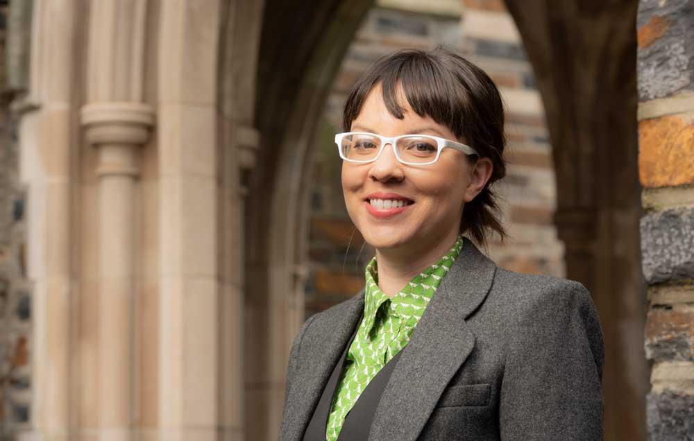 A headshot of Jessica Stark, who is wearing a green button up, gray blazer and white framed glasses.