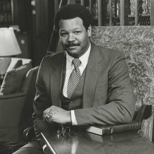 A black-and-white photo of Clarence Newsome posing with his arm on a book as he sits.